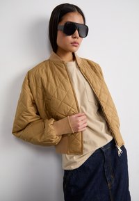 Chaqueta bomber acolchada beige con puños de canalé, combinada con una camiseta beige clara y una falda de mezclilla oscura, complementada con gafas de sol negras de gran tamaño.
