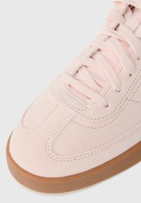 Adidași din suede roz deschis, cu detalii cusute, șireturi plate și o talpă din cauciuc texturată. Vârf rotunjit și suprafață netedă.