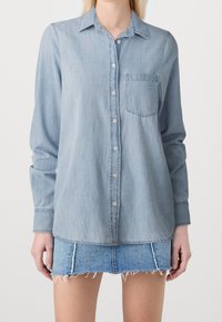 Woman wearing light blue button-up denim shirt and frayed hem denim mini skirt, standing against plain background.