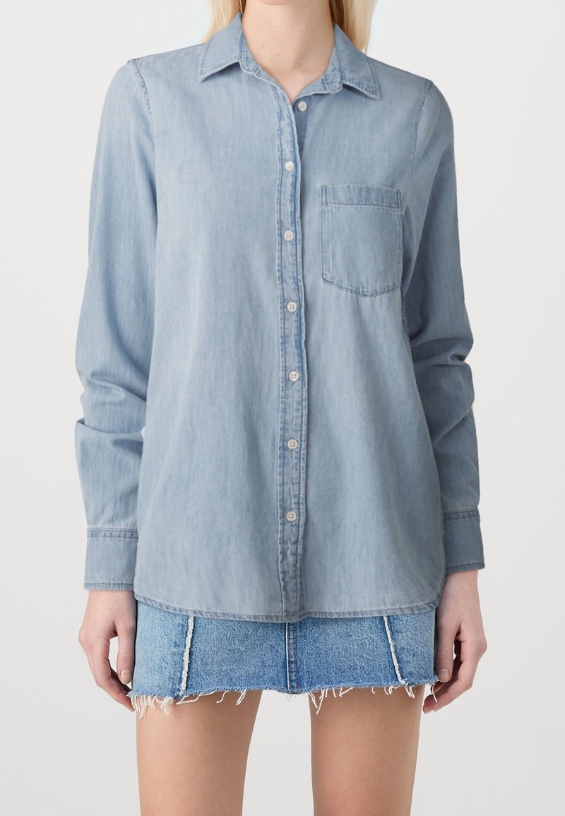 Woman wearing light blue button-up denim shirt and frayed hem denim mini skirt, standing against plain background.