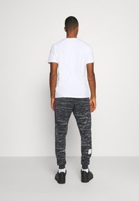 Svarta joggers med kamouflagemönster och sidosträck samt elastiska muddar, i kombination med en vit t-shirt med kort ärm. Stående framför en enkel bakgrund.