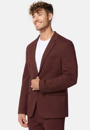 Bordeauxfarbener Blazer aus glattem Stoff, mit Reverskragen, einer Brusttasche und geknöpften Manschetten. Über einem weißen T-Shirt getragen.