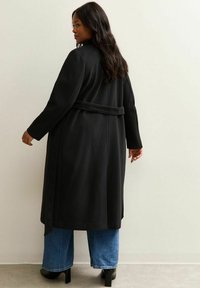 Zwarte wollen jas met lange mouwen, een ceintuur in de taille en een losse pasvorm, gecombineerd met wijde blauwe spijkerbroeken en zwarte hakken.
