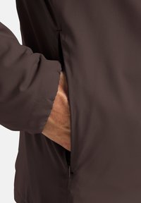 Veste légère marron avec une surface texturée, dotée d'une poche latérale et de poignets élastiques, conçue pour le confort et la liberté de mouvement.