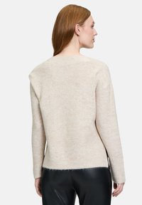 Beige gestrickter Pullover mit strukturierter Oberfläche, mit weitem Ausschnitt und geripptem Saum, getragen über einer schwarzen Hose.