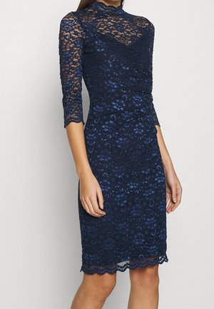 Granatowa koronkowa sukienka z długimi rękawami, wysokim dekoltem, dopasowaną sylwetką, kwiatowym wzorem i falbankowanym dołem. Materiał ma fakturowany wygląd.
