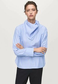 Young woman with folded arms wearing light blue and white striped long-sleeve shirt with a draped collar and black pants against a plain background.