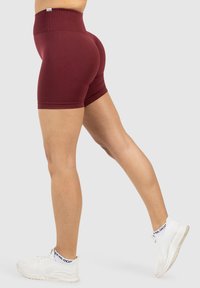 Bordowe szorty z wysokim stanem, wykonane z elastycznego materiału, o dopasowanym kroju i gładkiej fakturze, w połączeniu z białymi butami sportowymi.