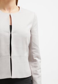 Veste gris clair structurée avec un motif géométrique, col rond, manches longues et fermeture éclair à l'avant.