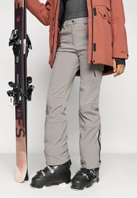 Suusavarustus sisaldab heledat halli kitsast lõikega pükst, millel on tõmblukuga taskud ja tekstuuriga viimistlus, mida täiendavad mustad suusasaapad ja lumelaud.