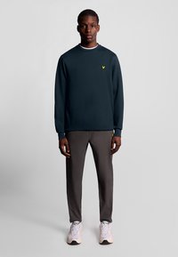 Mörkblå sweatshirt med lång ärm och en liten gul logotyp på bröstet, matchad med grå slim-fit byxor och vita sneakers.