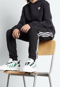 Svart hoodies med vitt Adidas-logga, svarta joggers med vita sidostrecker och vita sneakers med gröna ränder och detaljer.