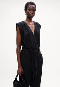 Mono negro con escote en V profundo, hombros almohadillados, cintura ceñida con cinturón y bolsillos laterales. La modelo sostiene un pequeño bolso negro. Textura de tela suave.