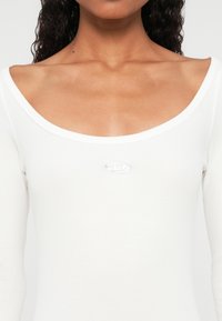 Közelkép egy nőről, aki fehér, kerek nyakkivágású, bordázott felsőt visel, melynek mellkasán középen egy kicsi, ovális, fémes logó található.