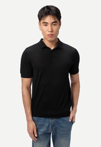 Jeune homme aux cheveux courts et foncés, portant un polo noir à manches courtes et un jean bleu, debout devant un fond blanc uni.