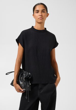 Femme portant un haut noir texturé à manches courtes et un pantalon noir tenant une pochette noire à clous et cordon dans une main.