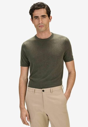 Man draagt een olijfgroene, korte mouw gebreide trui die in beige broek is gestopt, staat met één hand in de broekzak tegen een witte achtergrond.
