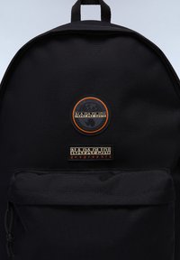 Svart tygryggsäck med texturerad yta, med två logotyper: en cirkulär patch med orange accenter och en rektangulär etikett. Enkel design.