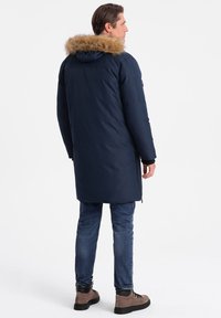 Námořnická modrá parka s kapucí s umělou kožešinou, dlouhými rukávy a bočními kapsami, nošená přes džíny a hnědé boty.