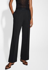 Pantalons noirs à jambes larges avec une finition lisse et texturée, dotés de poches latérales et d'une taille ajustée, complétés par des sandales noires à lanières.