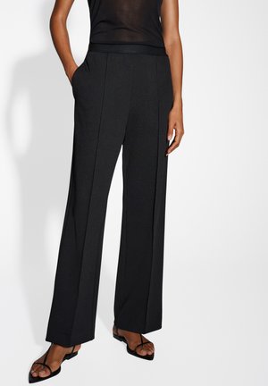 Schwarze, weite Hosen mit einer glatten, strukturierten Oberfläche, ausgestattet mit Seitenaschen und einem maßgeschneiderten Bund, ergänzt durch schwarze, geschlossene Sandalen mit Riemchen.