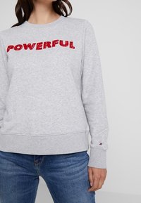 Sudadera gris con el texto rojo "PODEROSO", cuello redondo, puños y dobladillo acanalados. Combinada con jeans azules que presentan un pequeño logo en la manga.