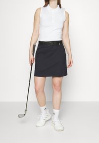 Biała koszulka bez rękawów polo, granatowa spódnica golfowa z plisami z przodu, czarny pasek, białe buty sportowe i kij golfowy w ręku.