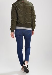 Zelená bomber bunda s rukávy s nařasením na zadní straně, pletenými manžetami a límcem, v kombinaci s tmavě modrými skinny džínami a černobílými teniskami.
