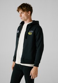 Crna zip-up dukserica s kapuljačom, rebrastim rukavima i donjim rubom. Sadrži bijeli i žuti izvezeni tekst i logotipe na prsima. Nosiva preko svijetle košulje.