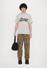 Gray cotton t-shirt with a black outlined leaping animal graphic and beige leopard print pants. Model holds a black bag and wears black shoes.