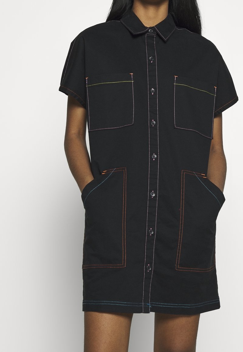 Robe noire à manches courtes avec un devant boutonné, présentant des coutures de couleur contrastante, deux grandes poches avant et un col de chemise.