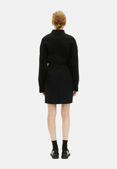 Zwarte gebreide trui met hoge kraag, gecombineerd met een zwarte mini-jurk. Model draagt zwarte sokken en glanzende schoenen, gezien vanuit de achterkant.