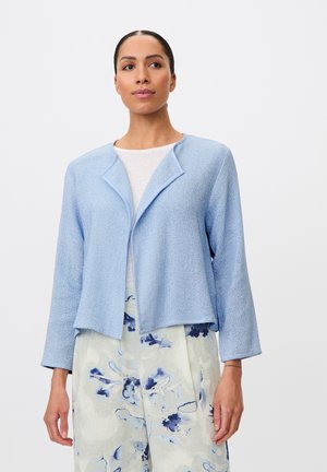 Kvinde iført en lyseblå åben-front tekstureret jakke over en hvid top og hvide bukser med blå blomsterprint, stående mod en ensfarvet baggrund.