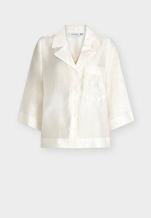 Chemise en soie crème à manches courtes avec un col cranté, une poche poitrine unique et des initiales brodées délicatement sur la poche.