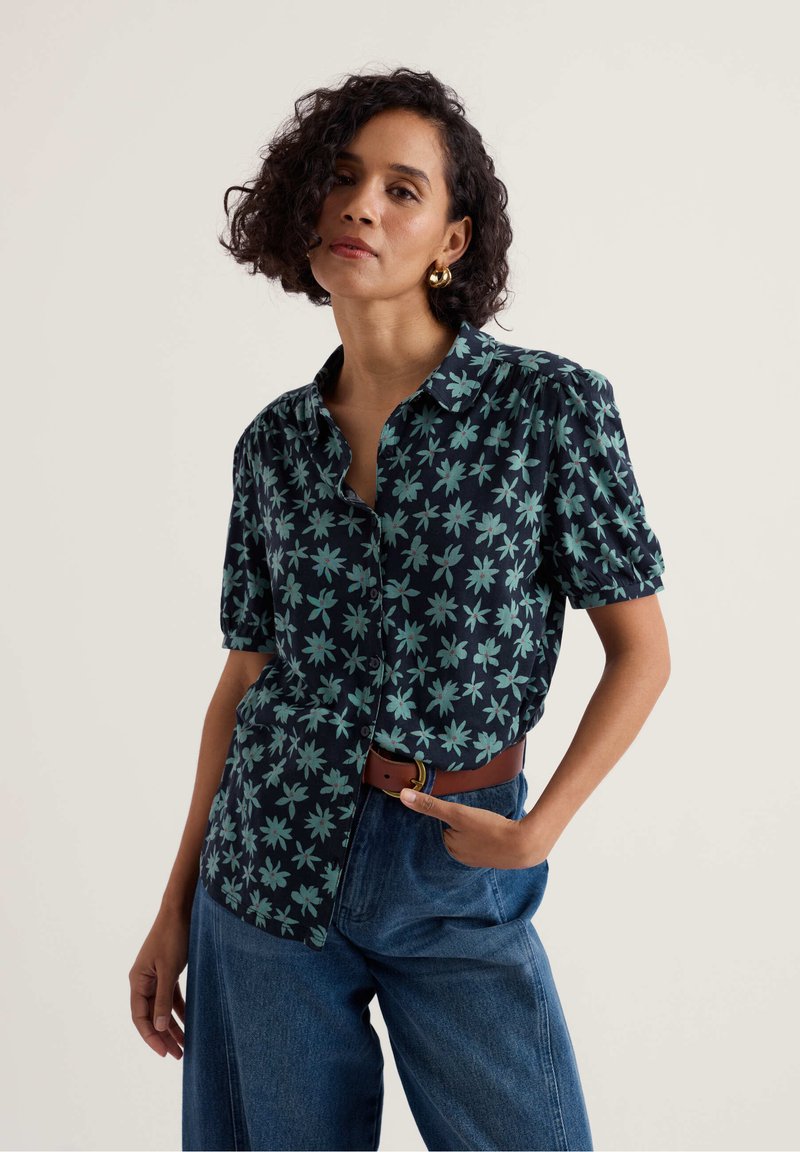 Frau mit dunklen lockigen Haaren, die eine blaue, kurzärmelige Bluse mit Blumenmuster und Knöpfen trägt, goldene Creolen, braunen Gürtel und blaue Jeans, steht mit einer Hand in der Tasche.