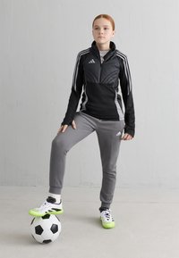 Melna un pelēka sporta uzvalka komplekts, kas sastāv no iešūta jakas, šaurām pelēkām biksēm un spilgti zaļām futbolas apaviem, stāv uz melna un balta futbolbumbas.