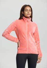 Vrouw in een koraalroze, gestructureerde fleece jack met rits, gekleed in zwarte broek, staand met één hand in haar zak tegen een effen achtergrond.