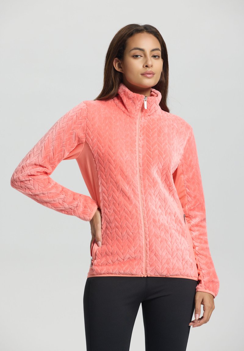 Vrouw in een koraalroze, gestructureerde fleece jack met rits, gekleed in zwarte broek, staand met één hand in haar zak tegen een effen achtergrond.