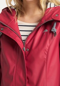 Czerwona wodoodporna kurtka z zamkiem błyskawicznym, zapięciem na napy i podszytą dzianiną kapturem. Posiada małe logo parasolki i kontrastowe paski podszewki.