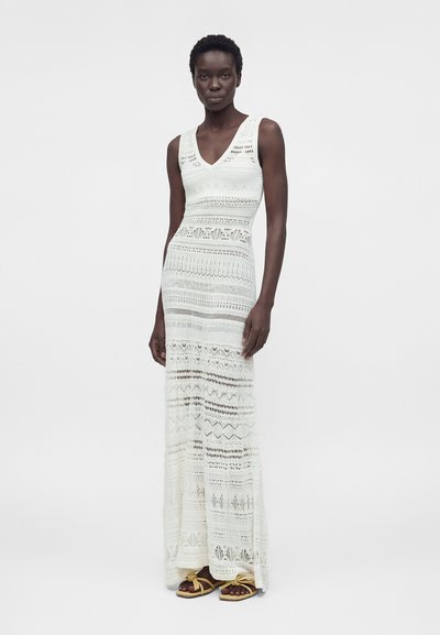 Λεπτή γυναίκα που φοράει λευκό αμάνικο πλεκτό μακρύ φόρεμα και κίτρινα σανδάλια, στέκεται μπροστά σε ένα απλό λευκό φόντο.