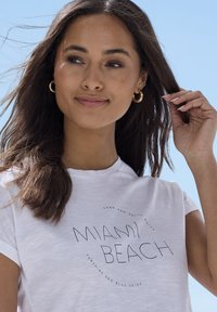 White t-shirt featuring a circular graphic with "MIAMI BEACH" and surrounding text. The fabric appears soft with a relaxed fit.