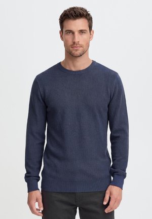 Homme aux cheveux courts bruns, portant un pull texturé bleu marine à manches longues et un pantalon noir, debout devant un fond blanc uni.