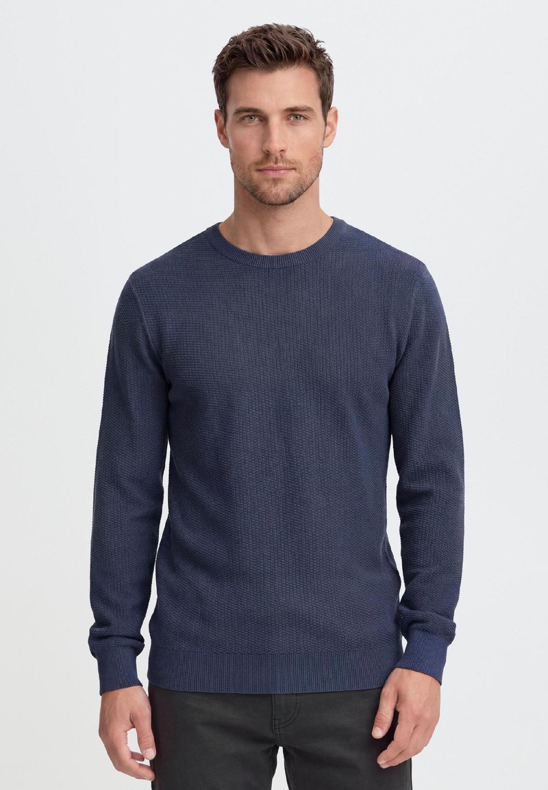 Uomo con capelli castani corti che indossa un maglione blu navy a manica lunga e pantaloni neri, in piedi davanti a uno sfondo bianco semplice.
