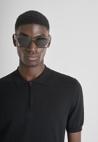 Crna polo majica s kratkim rukavima i ovratnikom na zatvarač, uparena s prozirnim, kutnim sunčanim naočalama s tamnim lećama.