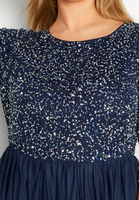 Námořnická modrá šaty se zdobeným korzetem a průsvitnou látkou. Mají kulatý výstřih, nařasený pas a splývavou sukni, což dodává texturu a lesk.