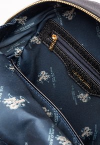 Interno di una borsa nera con cerniera, rivestito in tessuto blu navy, con un motivo di loghi chiari e una piccola tasca con cerniera in materiale nero testurizzato.