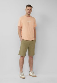 Lichtoranje t-shirt met ronde hals en logo, gecombineerd met olijfgroene knielange shorts en beige sneakers met witte accenten.