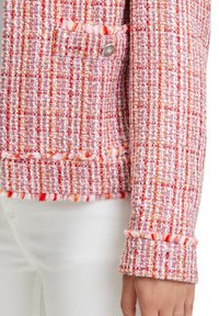 Różowa marynarka z tweedu z teksturowanym splotem i czerwonymi oraz pomarańczowymi akcentami. Srebrny detal guzika przy kieszeni. Frędzlowe wykończenie u dołu i na mankietach.