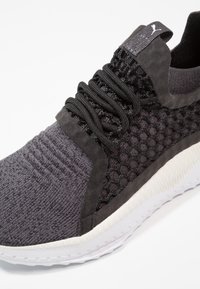 Chaussure de sport tricotée noire et gris foncé avec panneaux latéraux texturés, lacets noirs et semelle blanche coussinée.