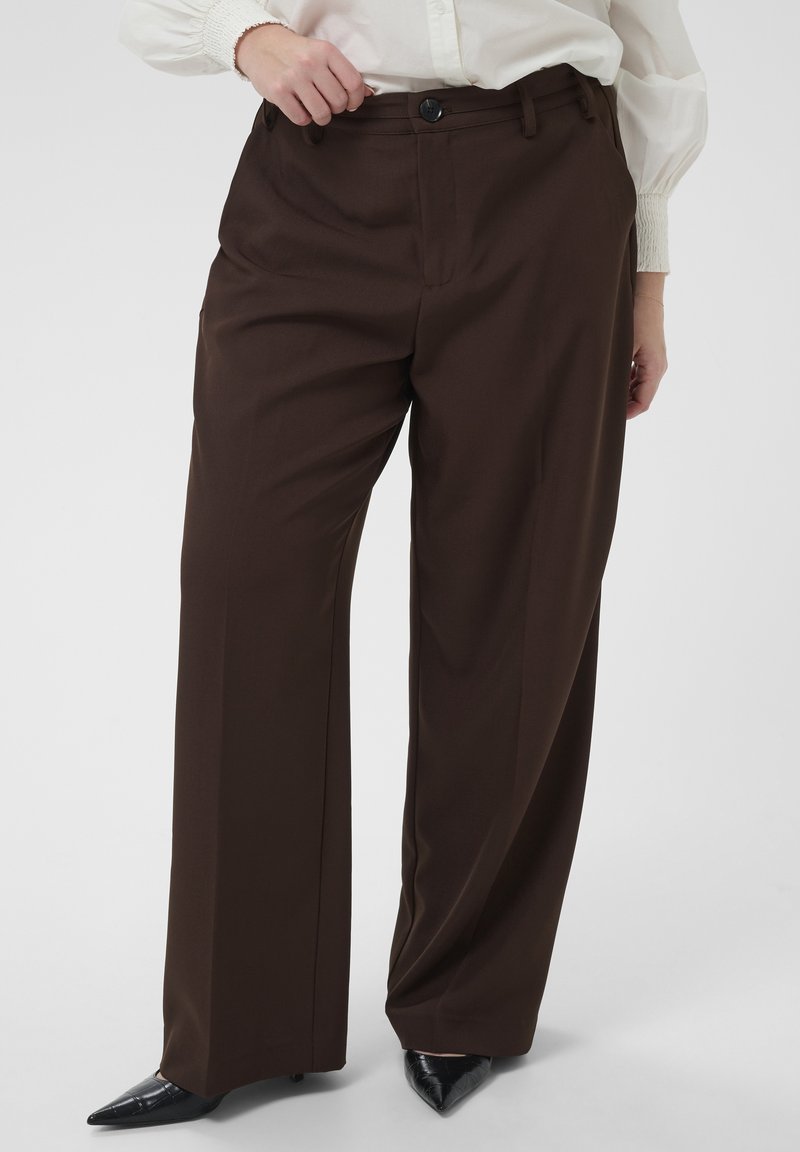 Femme portant un pantalon large marron foncé, un chemisier blanc et des talons pointus noirs, debout devant un fond uni.